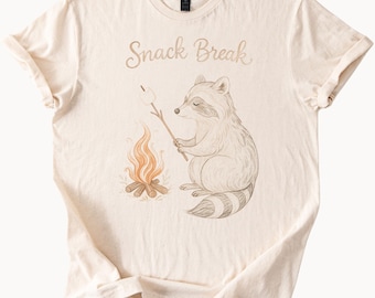 May include: A beige t-shirt with a raccoon illustration roasting a marshmallow over a campfire. The text "Snack Break" is printed on the shirt.