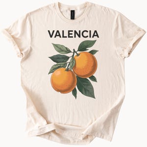 May include: A beige t-shirt with the word "VALENCIA" printed in black on the front. A branch with two orange oranges and green leaves is printed below the text.
