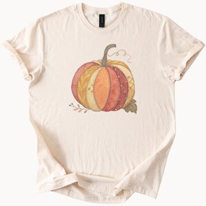 Puede incluir: Camiseta color crema con un diseño de calabaza en acuarela. La calabaza está segmentada con varios patrones y colores, incluyendo naranja, rojo y amarillo. Un tallo marrón y una hoja verde completan el gráfico temático de otoño.
