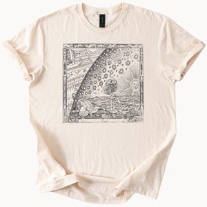 May include: A beige t-shirt with a black and white vintage illustration of a celestial map with a person lying down and a tree in the foreground.