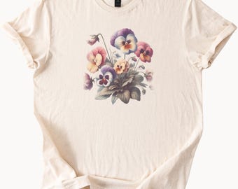 May include: Cream-colored t-shirt featuring a watercolor illustration of pansies in shades of purple, pink, and yellow. The floral design is centered on the shirt, with green leaves and stems. The shirt has short sleeves and a crew neck.