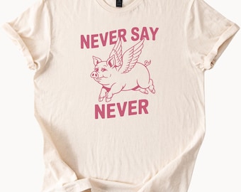 May include: A beige t-shirt with a pink pig with wings flying and the text "NEVER SAY NEVER" printed on it.