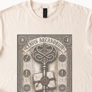 May include: A cream-colored t-shirt featuring a detailed dark brown graphic print of a large key, a sealed envelope, and the text CLAVIS ARCANORUM. The design includes intricate borders, circular symbols, and celestial motifs like a sun and moon.