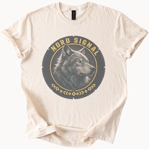 May include: A cream-colored short-sleeve t-shirt featuring a circular graphic on the chest. The graphic contains a detailed illustration of a wolf head in profile, the text NORD SIGNAL in gold, and a decorative gold geometric pattern at the bottom.