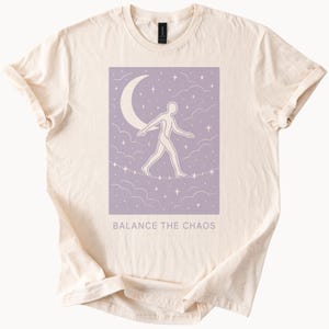 Puede incluir: Una camiseta beige con un gráfico morado de una persona caminando sobre una cuerda floja con una luna creciente sobre ella. El gráfico está rodeado de estrellas y nubes. El texto "Balance the Chaos" está impreso debajo del gráfico.