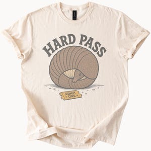 May include: A cream-colored t-shirt featuring a graphic of a brown armadillo curled into a ball. Above the armadillo, the text reads HARD PASS in dark gray. Below the armadillo, a yellow ticket is printed with the text ADMIT ONE in black ink.