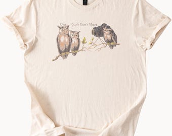 May include: Cream-colored t-shirt featuring a whimsical illustration of three owls perched on a branch. One owl is taking a photo with a vintage camera. The text "Owl Right Don't Move" is above the owls.