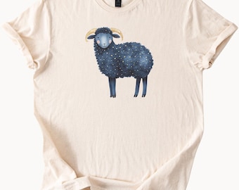 Puede incluir: Una camiseta de color crema con una oveja azul con estrellas doradas en su lana. La oveja tiene la nariz rosa y los ojos negros.