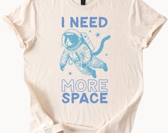 May include: A cream-colored t-shirt with a blue graphic of a cat wearing a space suit and the text "I NEED MORE SPACE".