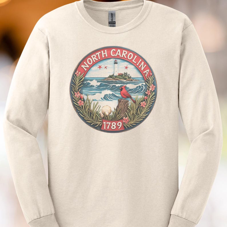 May include: Cream-colored long-sleeve shirt featuring a circular graphic of North Carolina. The graphic includes a lighthouse, ocean waves, a red cardinal, and the text "NORTH CAROLINA" and "1789".