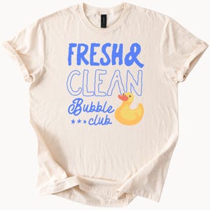May include: Cream-colored t-shirt with the text "FRESH & CLEAN Bubble club" in blue. A yellow rubber duck with an orange beak is featured below the text. The shirt has rolled-up sleeves and a crew neck.