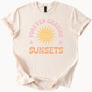 May include: A beige t-shirt with a pink and orange graphic design that says "Forever Chasing Sunsets" with a cartoon sun in the middle.