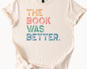 Pode incluir: Camiseta creme com o texto "THE BOOK WAS BETTER" em letras de bloco laranja, rosa, verde e azul. A camiseta tem mangas curtas e gola redonda.