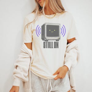 May include: A cream-colored short-sleeve t-shirt featuring a graphic of a gray vintage television set with a simple face, two yellow eyes, and a flat mouth. Purple signal waves flank the television, and the text STILL ON appears in gray below.
