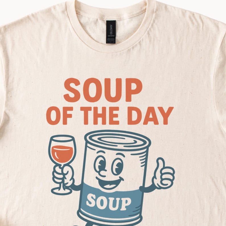 Op de afbeelding: Cr&egrave;mekleurig T-shirt met de tekst "SOUP OF THE DAY" in oranje. Daaronder een cartoon soepblik karakter met een wijnglas en een duim omhoog. Het soepblik heeft een blauw label met het woord "SOUP".