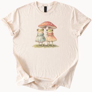 Può includere: T-shirt color crema con un'illustrazione stravagante di due rane antropomorfe sotto un fungo. Le rane indossano abiti blu e pesca, in piedi sull'erba. Il fungo ha un cappello rosso con pois bianchi.