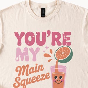 K&ouml;nnte beinhalten: Cremefarbenes T-Shirt mit dem Text "YOU'RE MY Main Squeeze" in Pink und Orange. Eine Grafik einer aufgeschnittenen Orange, die Saft in ein Glas mit einem l&auml;chelnden Gesicht gie&szlig;t, ist ebenfalls abgebildet.