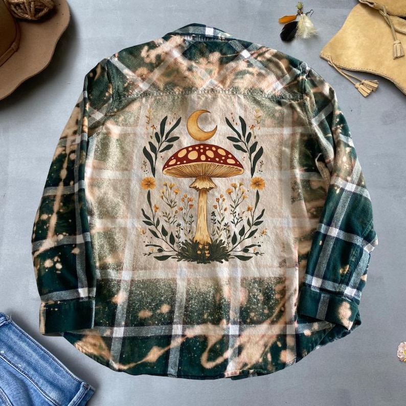 May include: A green and white plaid flannel shirt with a bleached effect. The back of the shirt features a mushroom with a crescent moon above it, surrounded by flowers and leaves.