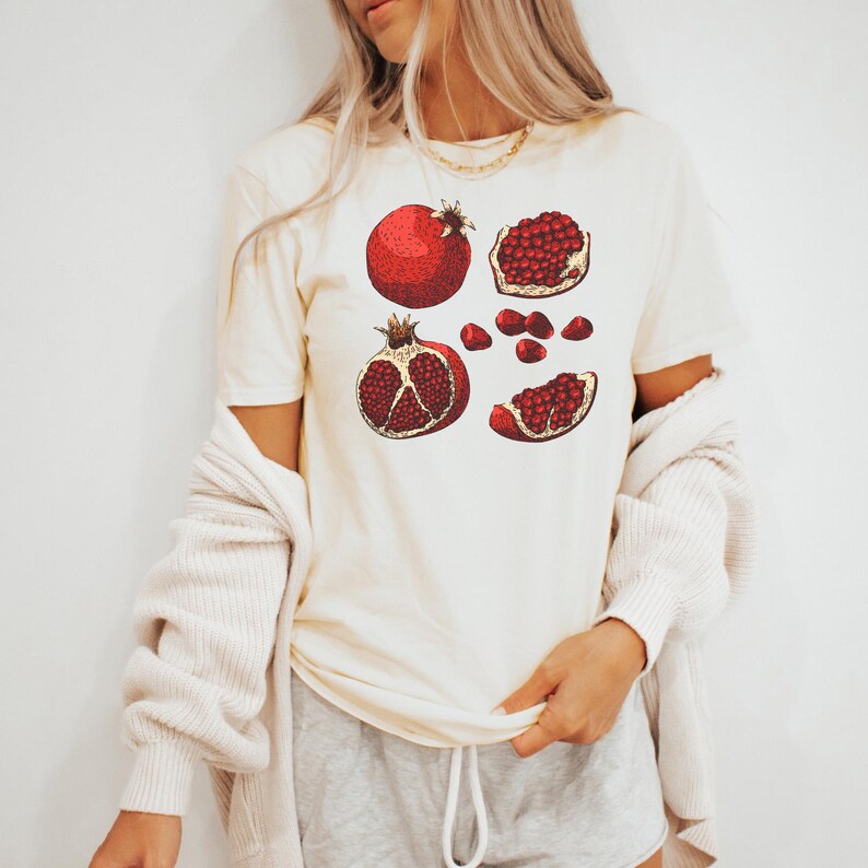 May include: Cream-colored t-shirt featuring a detailed illustration of pomegranates. The design includes whole and sliced pomegranates in shades of red and brown. The shirt is a short-sleeved, casual style.