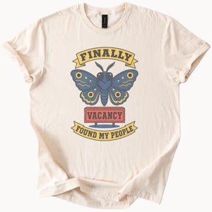 May include: A cream-colored short-sleeve t-shirt featuring a graphic of a blue moth above a red rectangular sign that reads VACANCY. Above the moth is a yellow banner reading FINALLY and below the sign is a yellow banner reading FOUND MY PEOPLE.