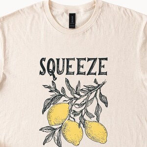 May include: A beige t-shirt with a black graphic of lemons on a branch and the word "SQUEEZE" in black lettering.