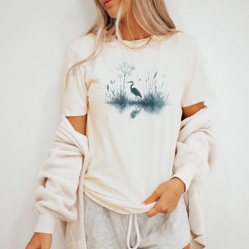 May include: A white t-shirt with a blue watercolor design of a heron standing in a marsh with tall grasses and reeds. The heron is facing left and its reflection is visible in the water.