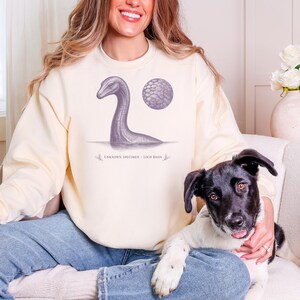 May include: A cream-colored crewneck sweatshirt featuring a dark gray graphic of a long-necked aquatic creature emerging from water, a circular textured detail, and the text "UNKNOWN SPECIMEN - LOCH BASIN" printed in a serif font below the illustration.