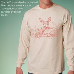 May include: A long-sleeved, beige shirt with a pink illustration of a doe and fawn. The text on the shirt reads: "Natural" is not dyed or bleached, The specks you see are soft natural fibres & tiny particles of cotton seeds.