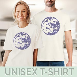 Op de afbeelding: Een wit unisex t-shirt met een paarse maanprint. De maan is een gestileerd ontwerp met een witte binnenkant en een paarse omtrek.