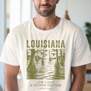 May include: Off-white t-shirt featuring a green graphic of a Louisiana swamp scene with trees and water. The word "LOUISIANA" is at the top, with "LOW LIGHT & DEEPER RHYTHM" and "SINCE 1812" below. The shirt is worn by a person.