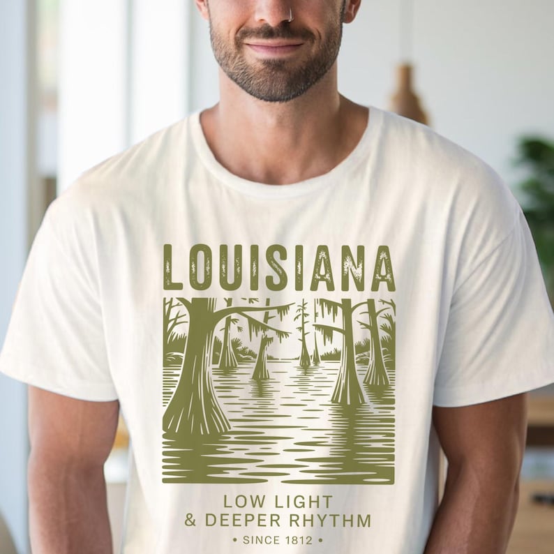 May include: Off-white t-shirt featuring a green graphic of a Louisiana swamp scene with trees and water. The word "LOUISIANA" is at the top, with "LOW LIGHT & DEEPER RHYTHM" and "SINCE 1812" below. The shirt is worn by a person.