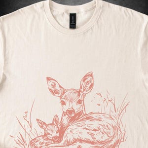 May include: Cream-coloured t-shirt with a coral-coloured illustration of a deer and a fawn in tall grass. The design is a detailed line drawing, with the deer's ears and eyes clearly visible. The shirt has a classic crew neck.