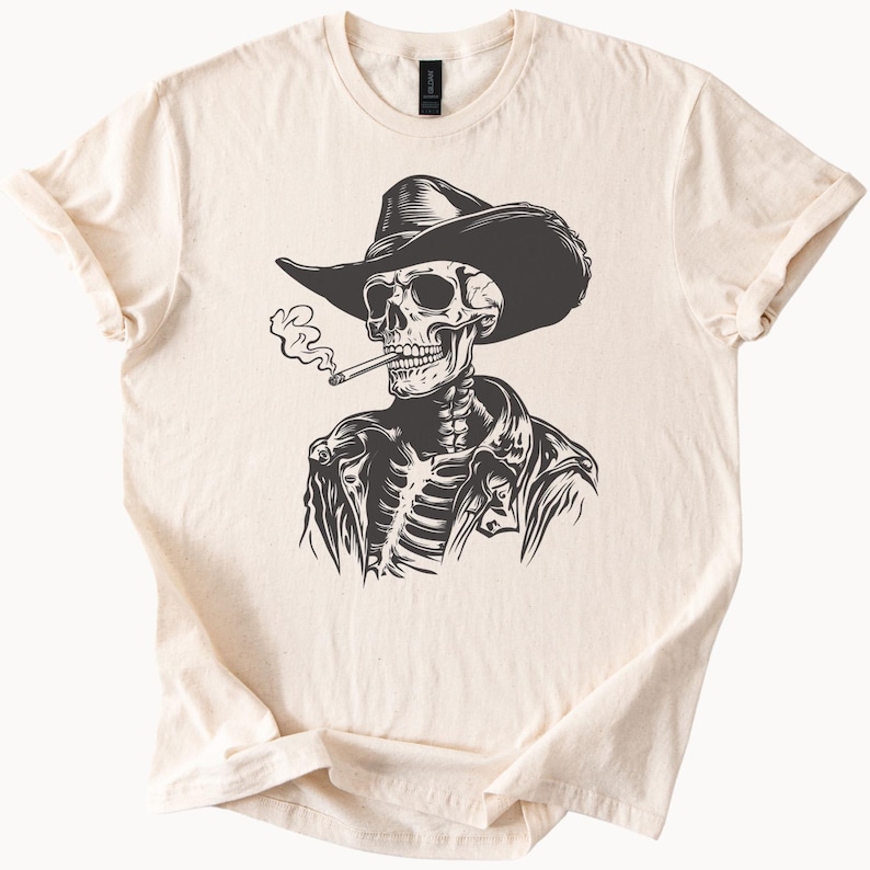 May include: A cream-coloured t-shirt with a black and white illustration of a skeleton wearing a cowboy hat and jacket, smoking a cigarette. The design is centered on the front of the shirt, with the sleeves rolled up.