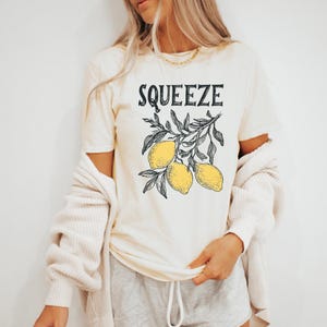 May include: A cream colored t-shirt with the text "Squeeze" in black lettering. The shirt features a graphic of a lemon tree branch with three yellow lemons.