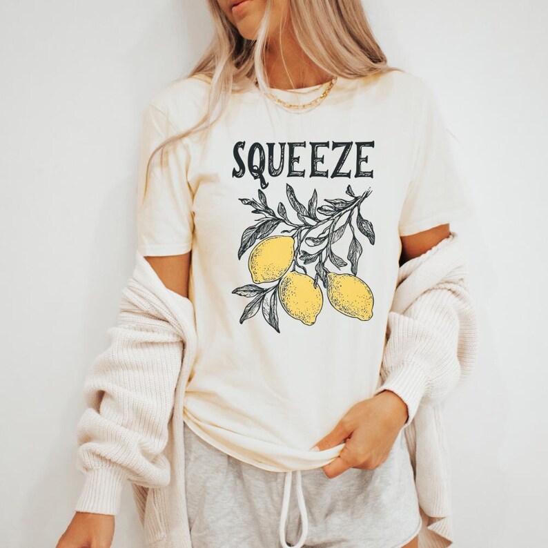 May include: A cream colored t-shirt with the text "Squeeze" in black lettering. The shirt features a graphic of a lemon tree branch with three yellow lemons.