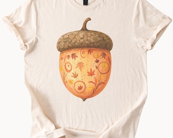 Puede incluir: Camiseta color crema con una gran ilustración detallada de una bellota. La bellota tiene una tapa marrón y un cuerpo naranja dorado adornado con hojas de otoño y motivos florales. La camiseta está hecha de material suave.