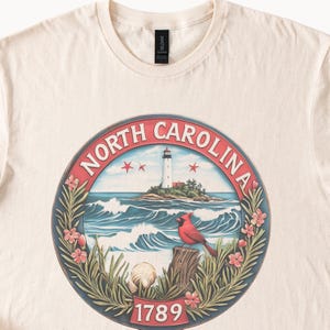 May include: Cream-colored t-shirt featuring a circular design with the words "NORTH CAROLINA" around the edge. The design includes a lighthouse, waves, a red cardinal, and the number "1789".