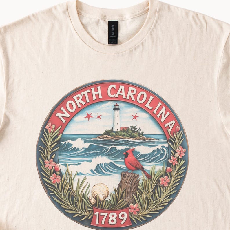 May include: Cream-colored t-shirt featuring a circular design with the words "NORTH CAROLINA" around the edge. The design includes a lighthouse, waves, a red cardinal, and the number "1789".