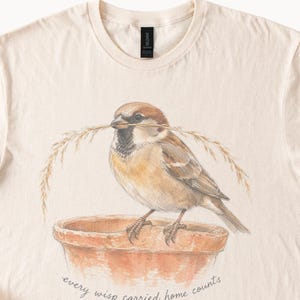 May include: A cream-colored t-shirt featuring a detailed illustration of a brown sparrow perched on a terracotta pot, holding a piece of dried grass in its beak. Below the pot, the text reads, every wisp carried home counts.