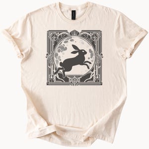 Puede incluir: Camiseta color crema con un gráfico en blanco y negro de un conejo saltando frente a una luna llena. La luna está enmarcada en un marco decorativo de estilo Art Déco. La camiseta tiene mangas cortas y cuello redondo.