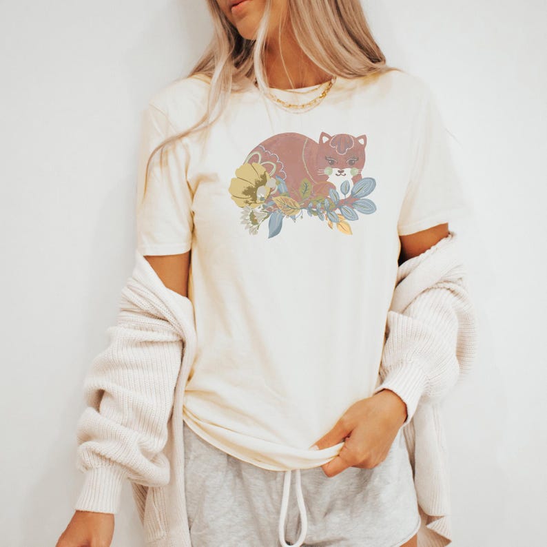 May include: Cream-coloured t-shirt with a stylised illustration of a resting cat surrounded by flowers and leaves in shades of brown, yellow, and blue. The graphic is centred on the front of the shirt. The shirt has short sleeves.