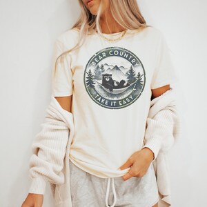以下が含まれることがあります： クリーム色のTシャツ。円形のグラフィックには、ハンモックでくつろぐクマ、山、木が描かれています。「Bear Country」と「Take it Easy」の文字はかすれたフォントです。半袖のシャツです。