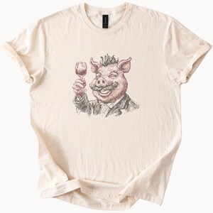Op de afbeelding: Crèmekleurig T-shirt met een humoristische illustratie van een varken met een snor, proostend met een glas rode wijn. Het varken draagt een colbert. De afbeelding is in rood, bruin en zwart.