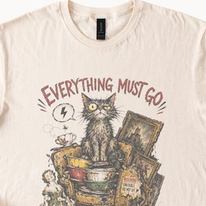 May include: A cream-colored t-shirt featuring a graphic of a disheveled cat sitting on a stack of bowls and furniture. The text "EVERYTHING MUST GO" is printed above the cat, and a small sign reads "WARNING UNSTABLE DO NOT SIT".
