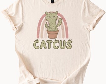 May include: A cream-coloured t-shirt with a quirky graphic. The design showcases a cactus shaped like a cat with a grumpy face, set against a pink and brown rainbow. The word "CATCUS" is printed below the image in a matching green colour.