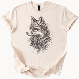 Op de afbeelding: Crèmekleurig T-shirt met een gedetailleerde zwart-wit illustratie van een vos. De kop en het bovenlichaam van de vos zijn afgebeeld met ingewikkelde patronen en krullen. Het shirt heeft een klassieke ronde hals en korte mouwen.