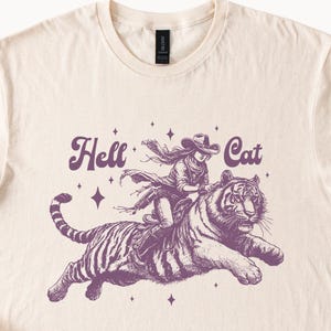 Könnte beinhalten: Ein weißes T-Shirt mit einer lilafarbenen Grafik einer Frau, die auf einem Tiger reitet. Der Text "Hell Cat" ist in einer Retro-Schriftart über der Grafik gedruckt.