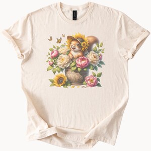 Puede incluir: Camiseta color crema con una ilustración caprichosa. Una ardilla con un sombrero de girasol está sentada en una maceta decorativa llena de rosas rosas y crema, girasoles y hojas verdes. Mariposas revolotean sobre las flores. La camiseta es de material suave.