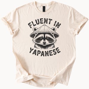 May include: Cream-colored t-shirt featuring a black and white illustration of a raccoon wearing a conical hat. The text "FLUENT IN YAPANESE" is printed above and below the raccoon image in a bold, sans-serif font. The shirt has rolled-up sleeves.