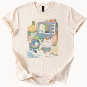 May include: A cream-colored t-shirt featuring a graphic illustration of a vintage blue television set displaying a color test pattern, surrounded by a bowl of blueberries, a dessert dish with whipped cream and a strawberry, bananas, and citrus.